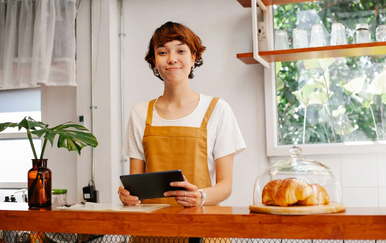 Café-Inhaberin mit Tablet hinter der Theke, daneben Croissants unter einer Glasglocke